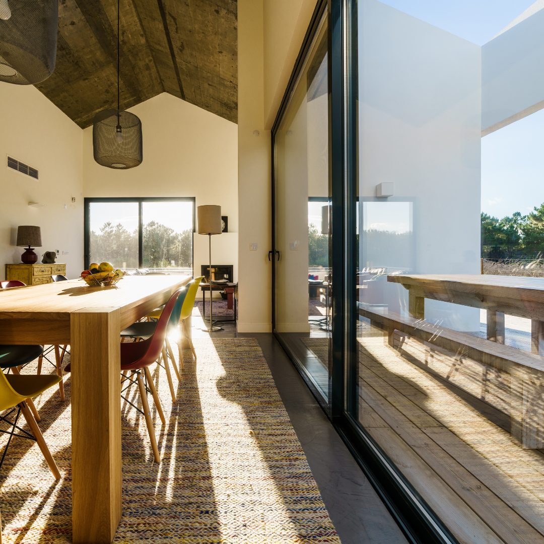 Sala de jantar iluminada com mesa de madeira, cadeiras coloridas e amplas portas de vidro com vista externa.

